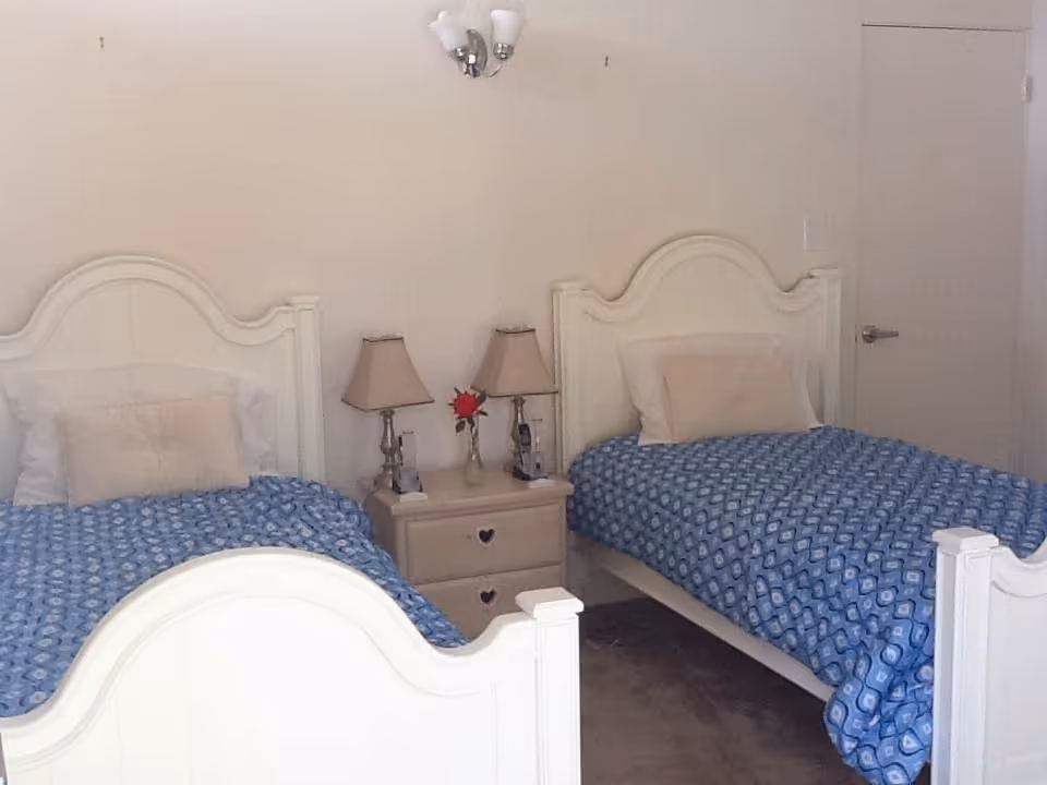 A bedroom with two white twin beds, each with a blue patterned bedspread and beige pillows. Between the beds is a small beige nightstand with two matching lamps and a vase with a red flower. The walls are light-colored, and there is a door visible on the right side of the room.