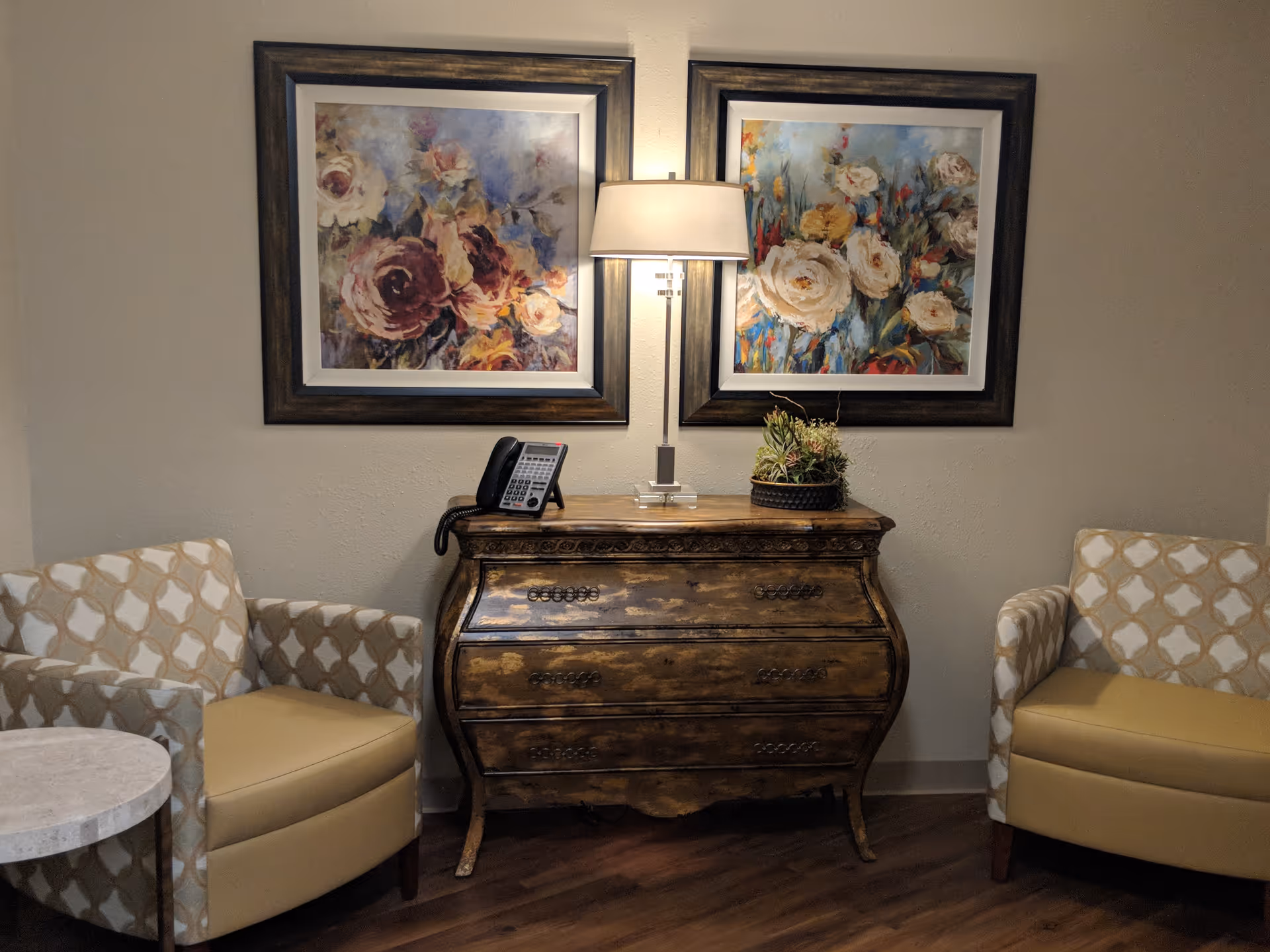 A cozy corner with two patterned armchairs facing each other, separated by a small round table on the left. Between the chairs is a vintage wooden chest with three drawers, on top of which sits a table lamp, a telephone, and a small plant arrangement. Above the chest are two framed floral paintings with dark wooden frames, illuminated by the lamp.