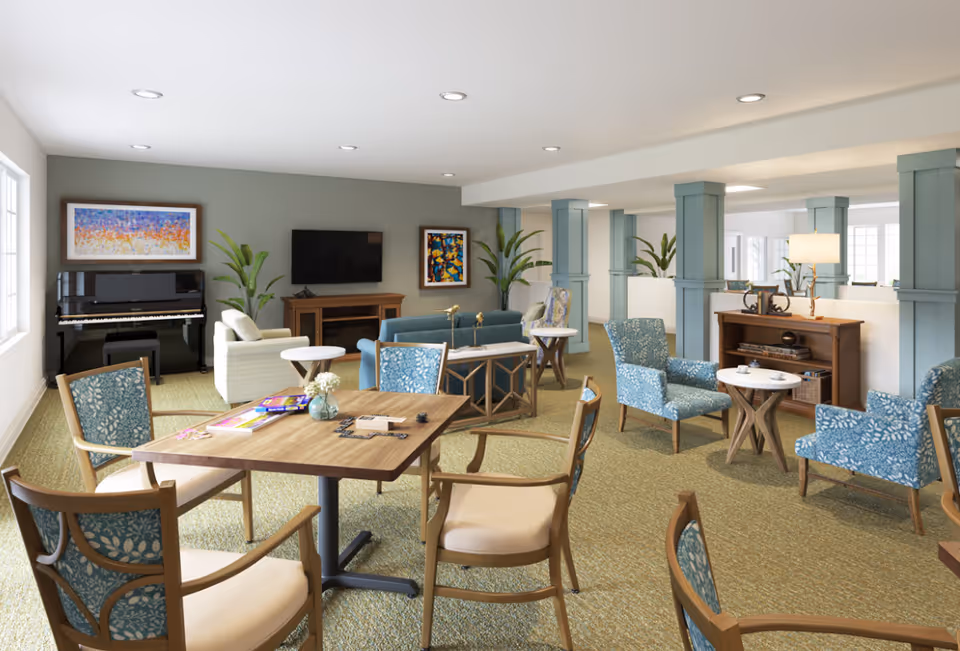 A spacious senior living common area with multiple seating arrangements including blue patterned chairs and beige cushioned chairs around wooden tables. There is a black upright piano against the wall, a flat-screen TV mounted on the wall above a wooden console, and colorful artwork on the walls. The room has green carpet, several potted plants, and light blue columns dividing the space. The area is well-lit with recessed ceiling lights and natural light from windows.