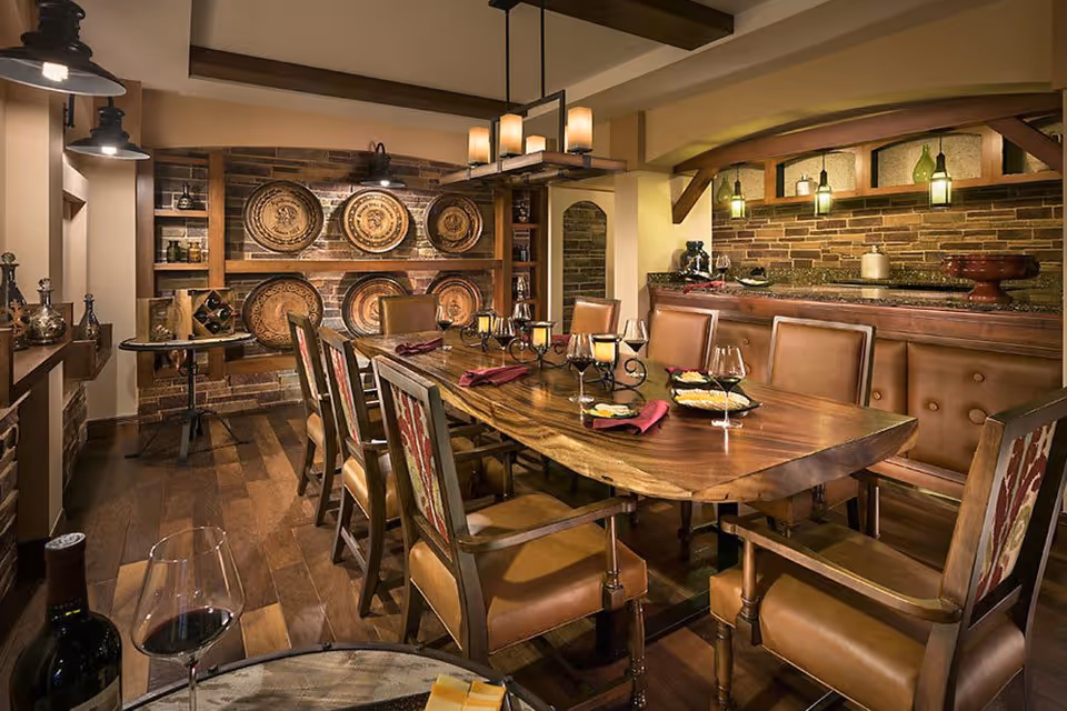 A warmly lit dining room with a long wooden table set with wine glasses, plates, and napkins. The room features leather chairs, a brick accent wall with decorative plates, wooden beams on the ceiling, and a counter area with hanging pendant lights.