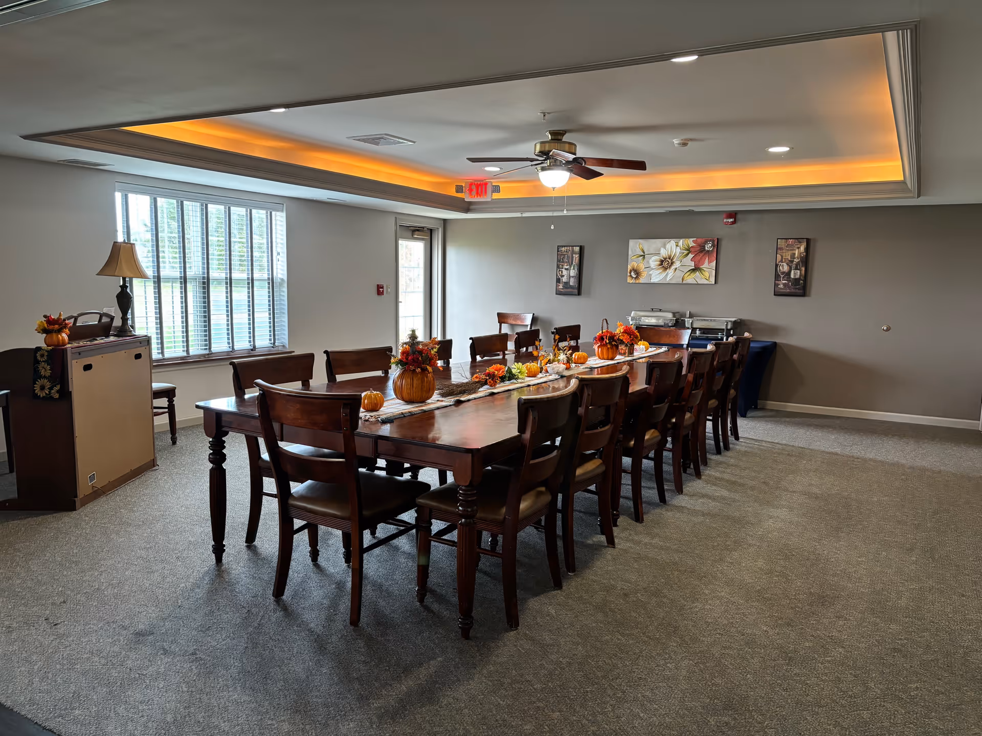 A dining room with a long wooden table surrounded by wooden chairs. The table is decorated with small pumpkins and autumn-themed floral arrangements. The room has a large window with blinds, a ceiling fan with a light, and soft recessed lighting along the ceiling edges. There are framed floral paintings on the wall and a serving station covered with a dark cloth in the corner.