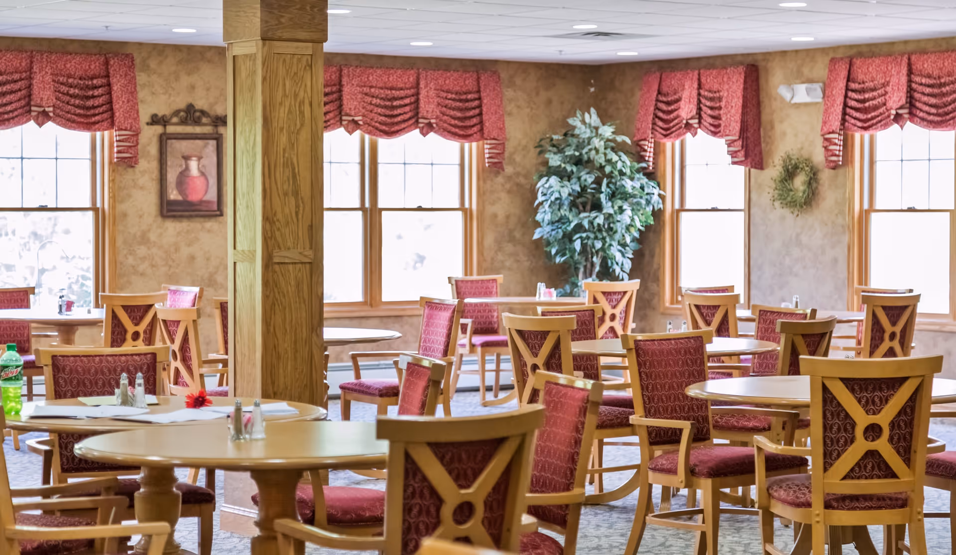 Sunlit dining room with round wooden tables and red upholstered chairs in a senior living community.