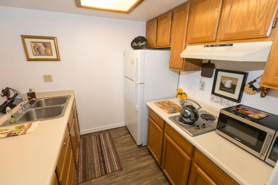 A compact kitchen with wooden cabinets, a white refrigerator, a double sink, a small electric stovetop with a kettle, a microwave, and various kitchen decorations including a framed mocha coffee picture and a small flower vase.