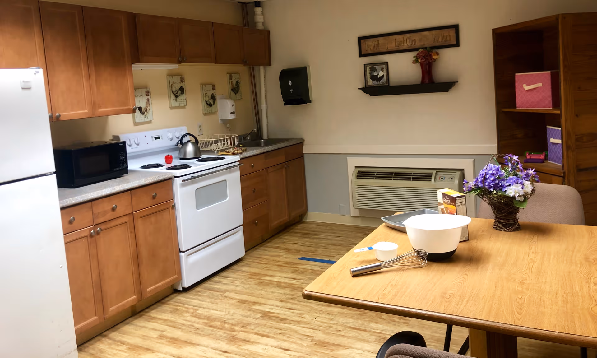 A kitchen area with wooden cabinets, a white refrigerator, a white stove with a kettle on it, and a microwave on the countertop. There is a sink with a dish rack and a paper towel dispenser on the wall. A wooden table with a whisk, mixing bowl, measuring cup, a box of baking mix, and a vase with purple flowers is in the foreground. A wooden shelving unit with colorful storage bins is visible in the background.