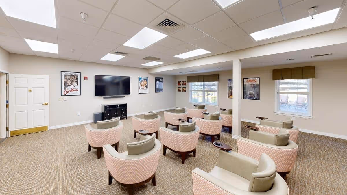A cozy common area with multiple cushioned armchairs arranged in rows facing a large flat-screen TV mounted on the wall. The room has beige walls, carpeted floor, and several movie posters including Casablanca and Jaws on the walls. There are windows with brown valances letting in natural light.