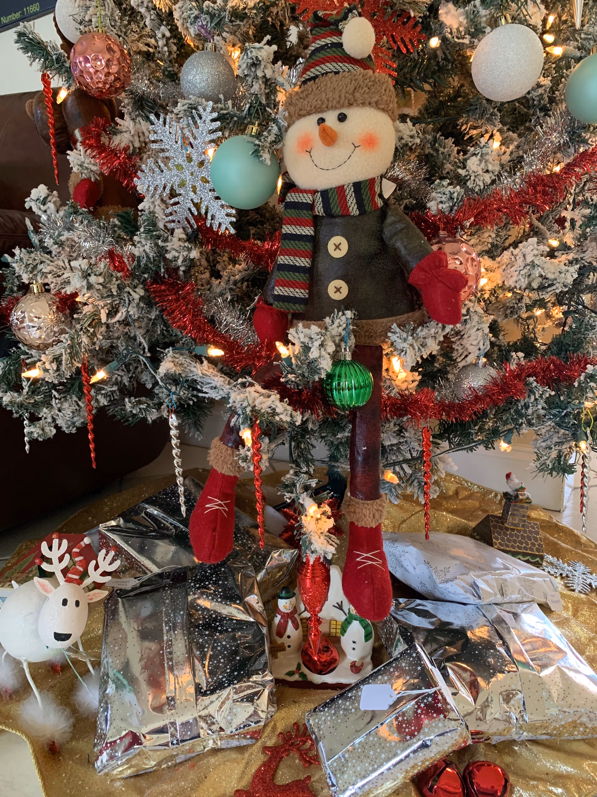 A decorated Christmas tree with snow-covered branches, red and silver tinsel, and various ornaments including a large smiling snowman figure wearing a striped scarf and red mittens. Under the tree are several shiny silver wrapped presents, a small white reindeer decoration, and a red vase with small festive figurines. The setting appears to be indoors with a brown couch partially visible in the background.