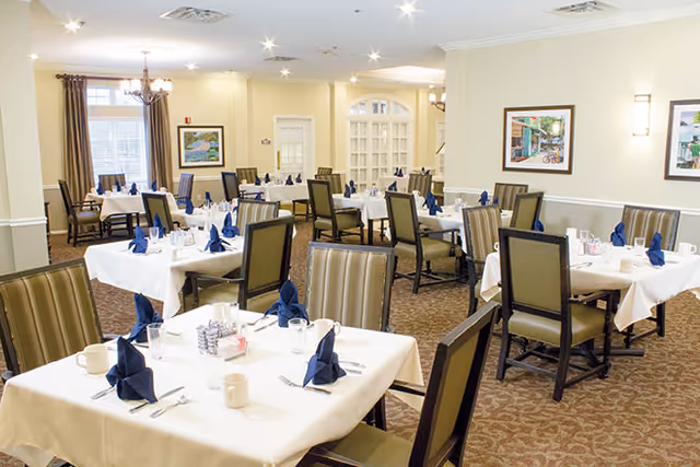 A bright dining room with multiple tables covered in white tablecloths, each set with blue folded napkins, glassware, and utensils. The room has beige walls, framed artwork, and large windows with curtains, creating a welcoming atmosphere.