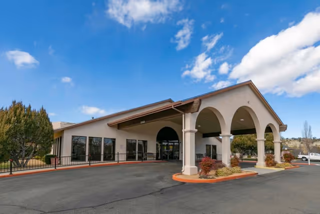 Exterior front view of a single-story building with a covered entrance supported by arches, surrounded by small landscaped bushes and trees under a partly cloudy blue sky.