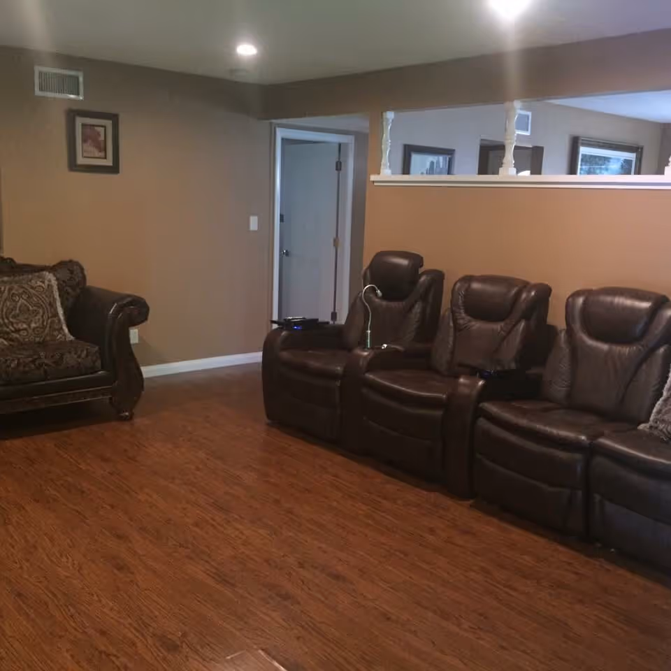 A living room with brown leather recliner chairs arranged in a row and a patterned fabric sofa against a tan wall. The floor is wooden, and there is a small half wall with white spindles separating the room from another area. A door and framed pictures are visible in the background.