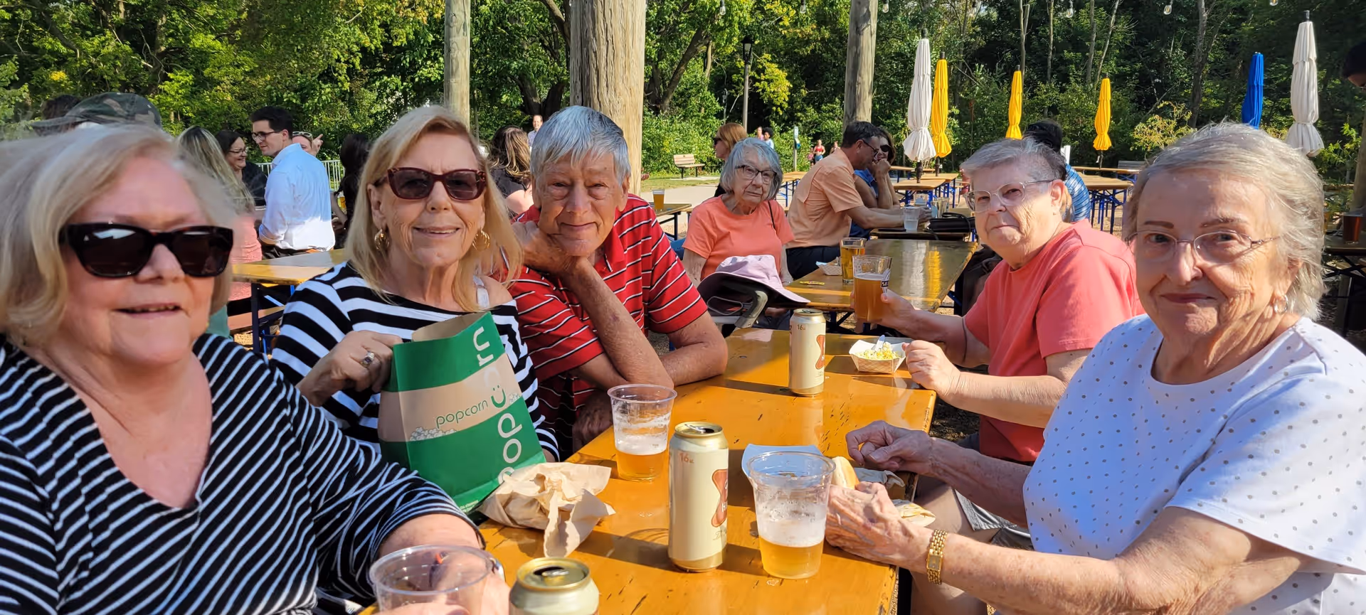 A group of elderly people sitting around a wooden picnic table outdoors, enjoying drinks and snacks on a sunny day. There are trees and umbrellas in the background, creating a relaxed and social atmosphere.