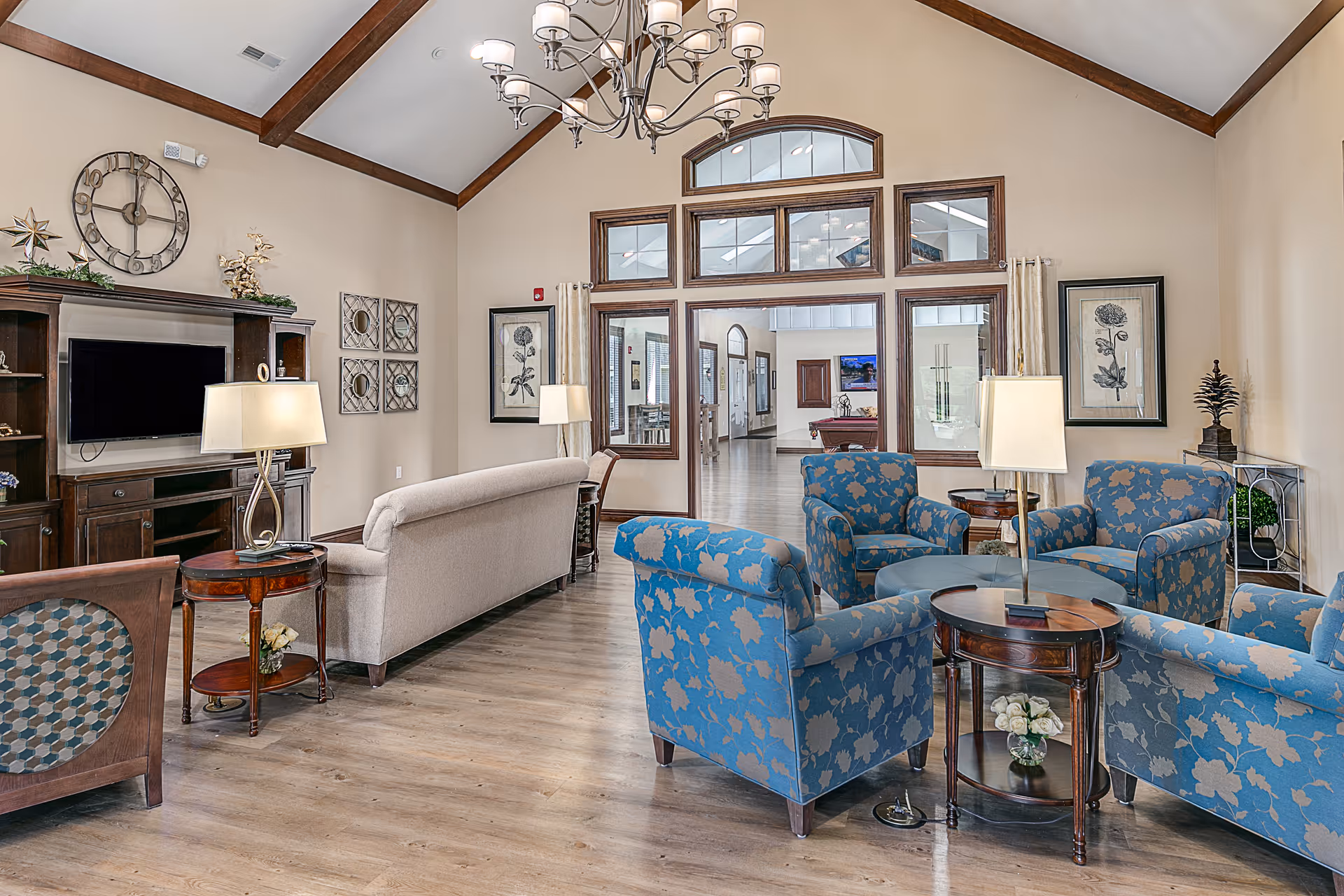 A spacious and well-lit living room area with high vaulted ceilings and wooden beams. The room features a beige sofa, four blue floral armchairs arranged around a circular coffee table, and several side tables with lamps. A large wooden entertainment center with a flat-screen TV is against one wall, decorated with plants and a large clock. The room has light wood flooring and large windows with wooden trim, allowing a view into an adjacent hallway and room.