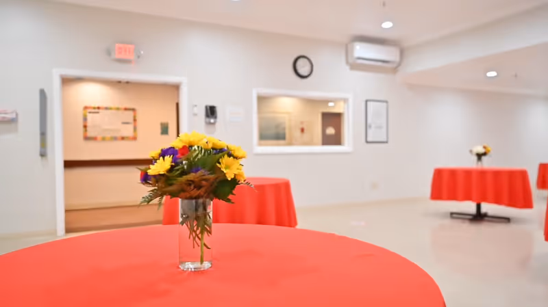 Interior room with round tables covered in red tablecloths, each table has a small vase with colorful flowers. The room has white walls, a clock, an air conditioning unit, and a window opening to another room.