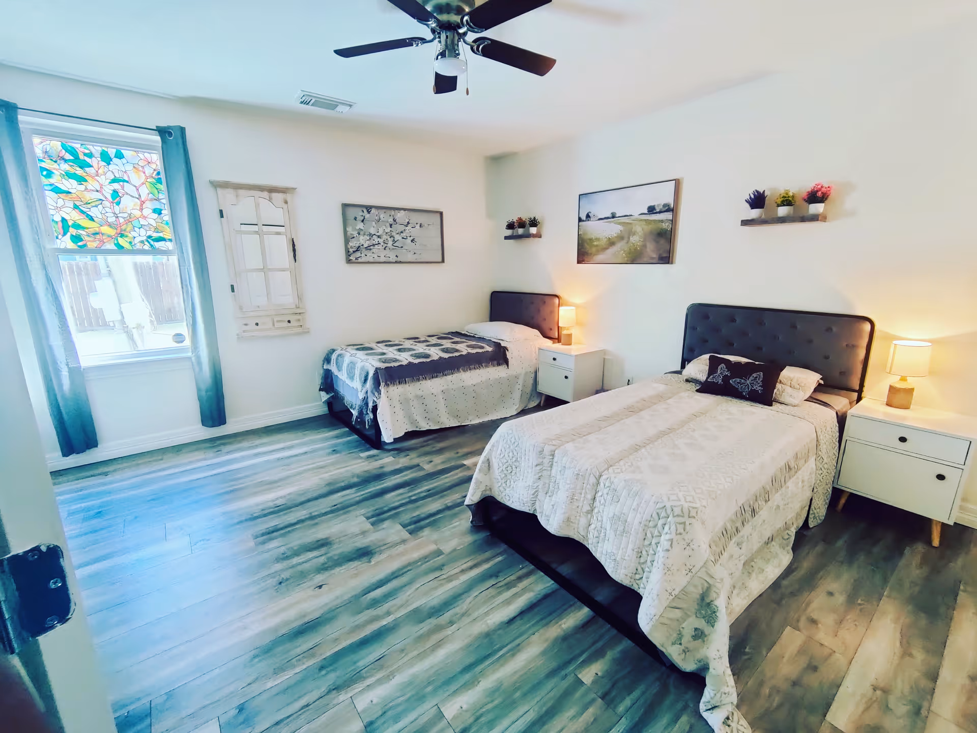 A bright bedroom with two twin beds, each with a tufted headboard and light-colored bedding. There are two white nightstands with lamps on either side of the beds. The room has light wood flooring, a ceiling fan, and a window with blue curtains and a colorful stained glass panel. The walls are decorated with framed artwork and small shelves holding potted plants.
