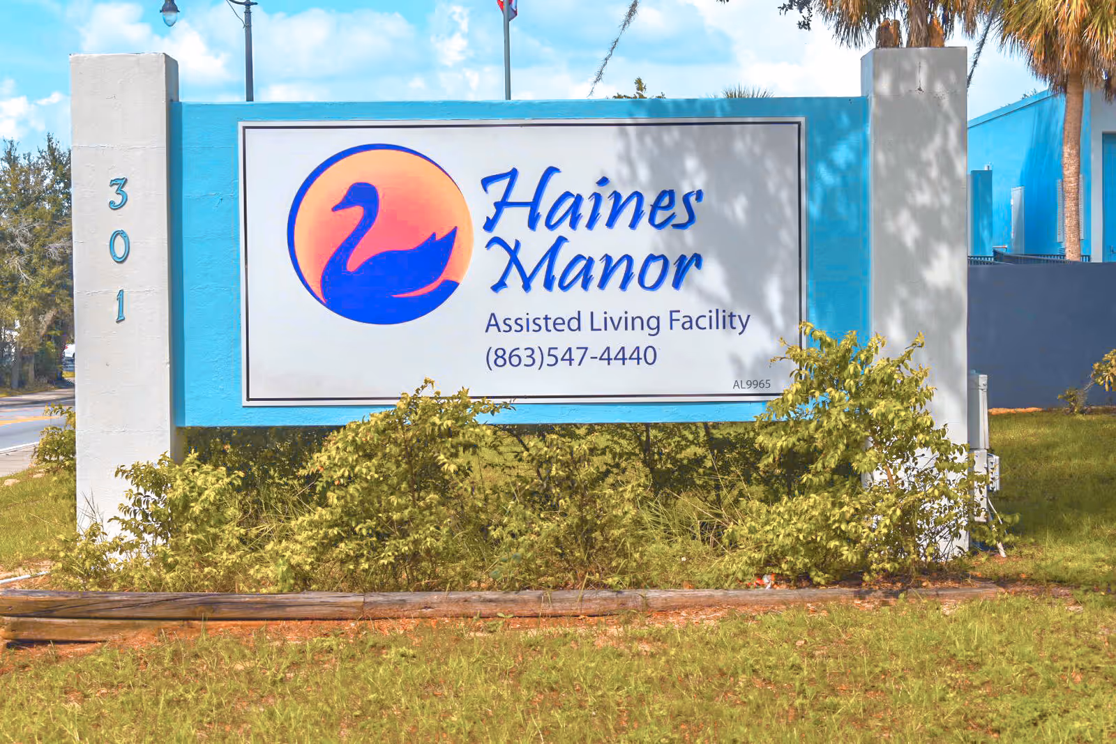 Outdoor sign for Haines Manor Assisted Living Facility with a blue swan logo, phone number, and address number 301 on a white and blue background surrounded by greenery.