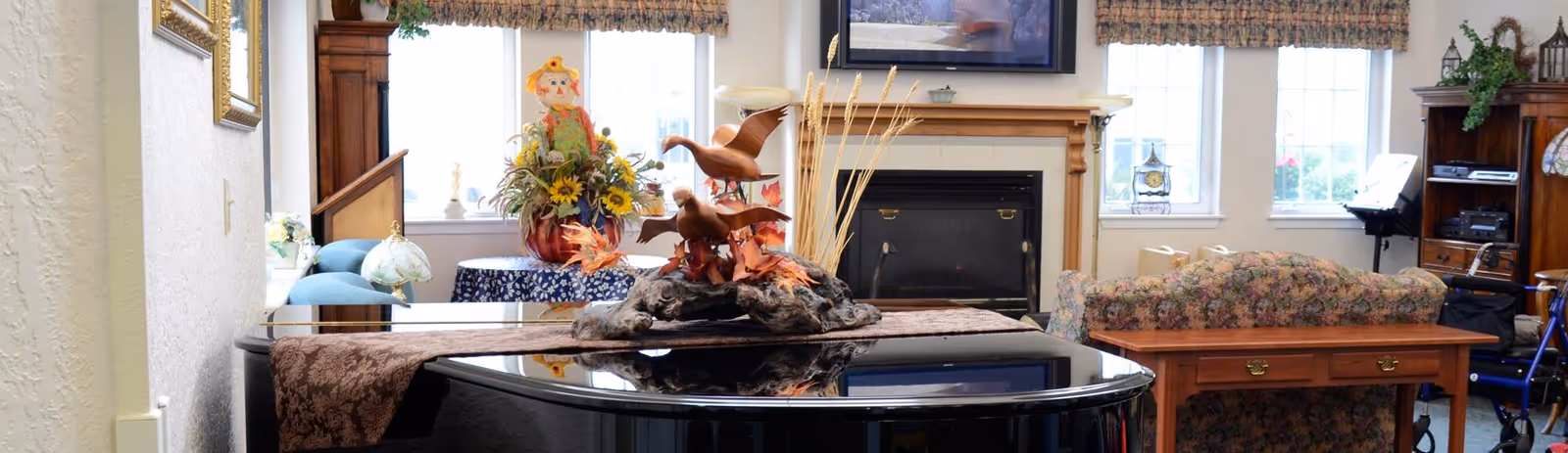 A cozy living room area featuring a black grand piano with a decorative centerpiece of wooden ducks and autumn leaves. Behind the piano, there is a floral-patterned couch with a wooden table, a fireplace with a wooden mantle, and windows letting in natural light. Additional decorations include a scarecrow figure with sunflowers on a small table and various lamps and framed pictures on the walls.