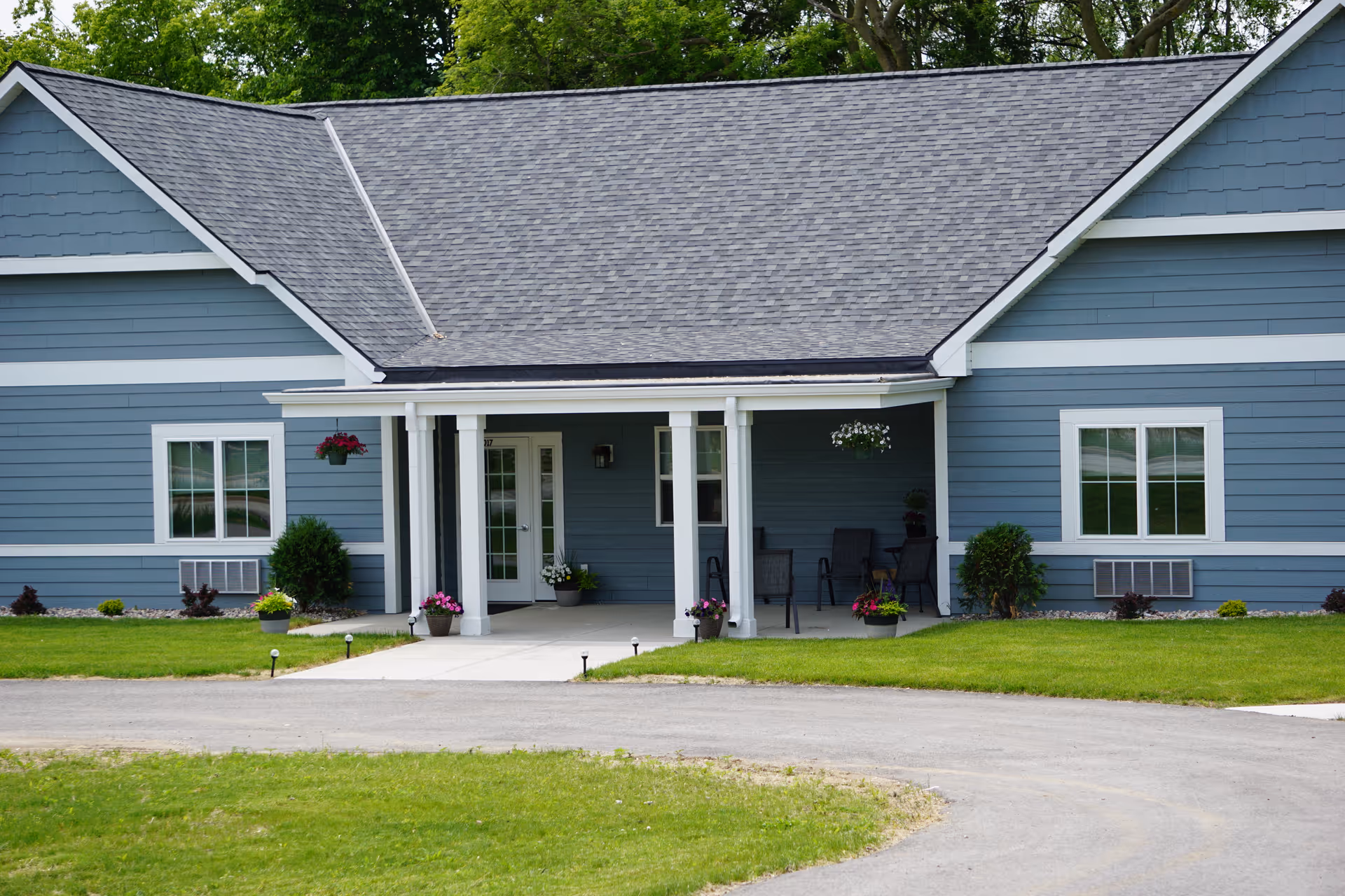 Front exterior view of a single-story blue senior living facility building with white trim, a covered porch with chairs and potted plants, and a well-maintained lawn and driveway.