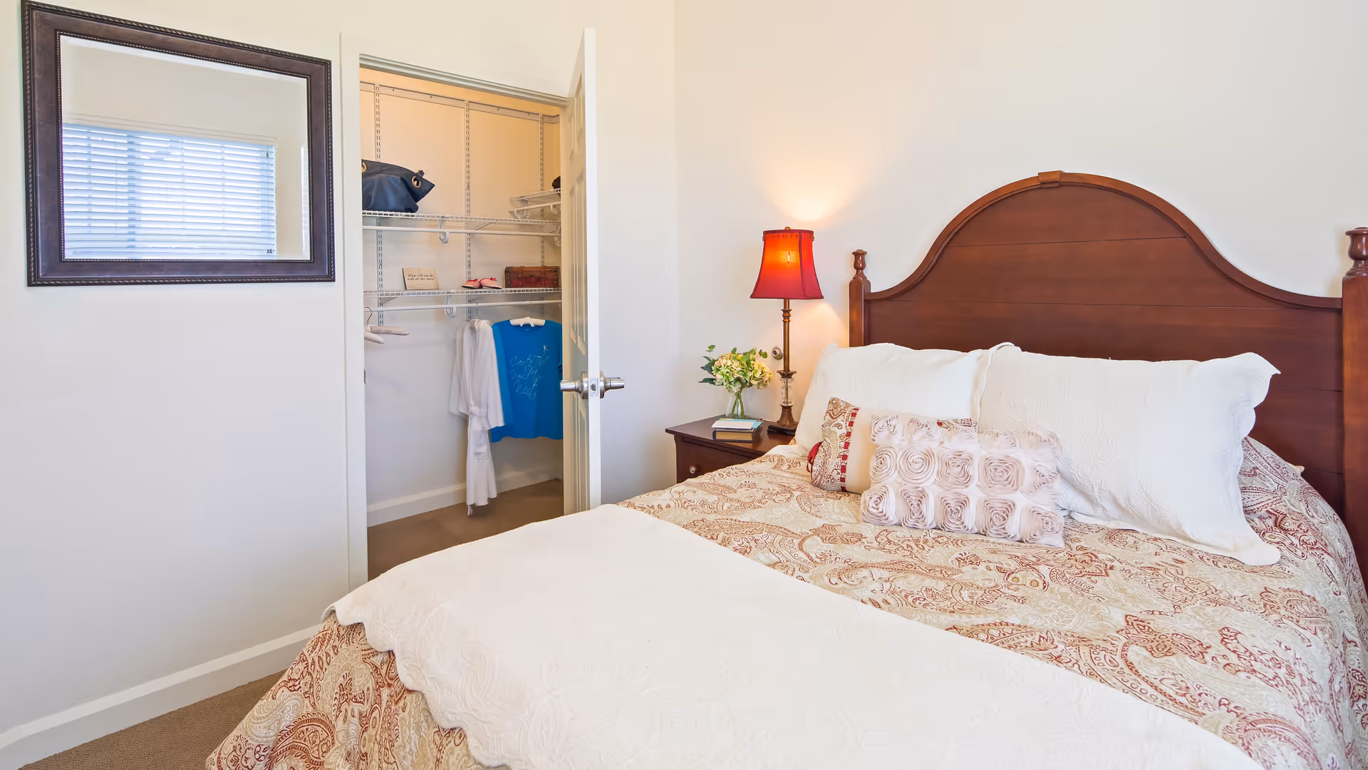 A bedroom with a wooden headboard bed dressed in patterned bedding and multiple pillows. A nightstand beside the bed holds a red lamp and a small vase with flowers. An open door reveals a walk-in closet with hanging clothes and shelves. A window with blinds and a framed mirror are on the wall opposite the bed.