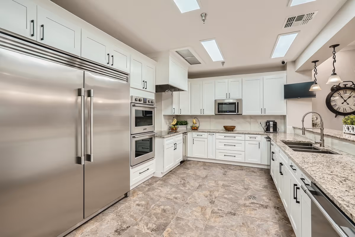 A modern kitchen with white cabinets, granite countertops, and stainless steel appliances including a large refrigerator, double oven, microwave, and dishwasher. The kitchen features a double sink with a high-arc faucet, pendant lighting, a wall clock, and a small TV mounted on the wall. The floor is tiled with a stone pattern.