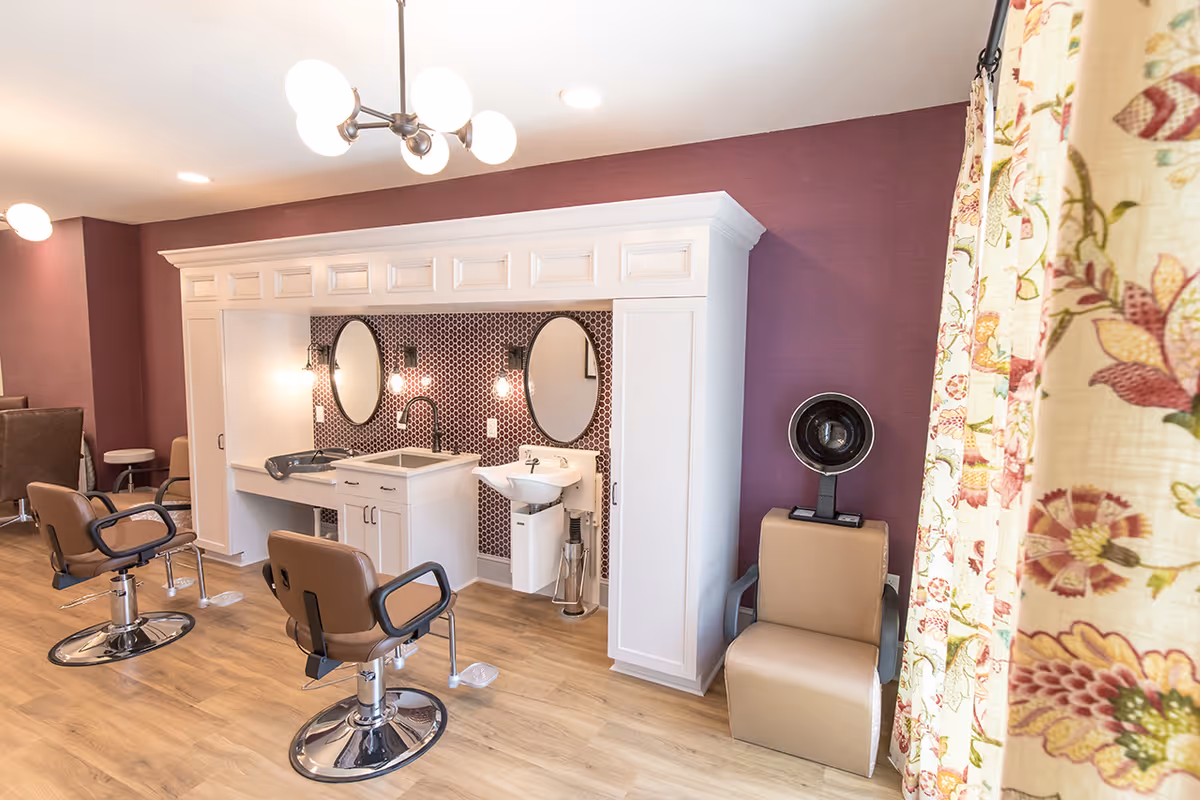 Interior view of a salon area with brown salon chairs, a white cabinet with two oval mirrors, sinks, and hair washing stations. The walls are painted purple, and there is a floral curtain on the right side.
