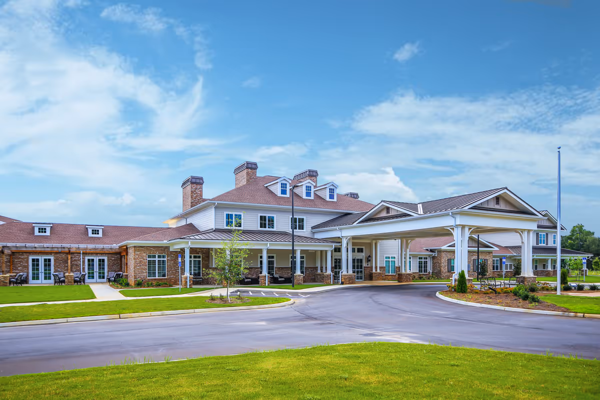 Exterior view of Grand South Senior Living facility showing a large building with a covered entrance, brick and siding facade, multiple windows, and a well-maintained lawn under a partly cloudy blue sky.