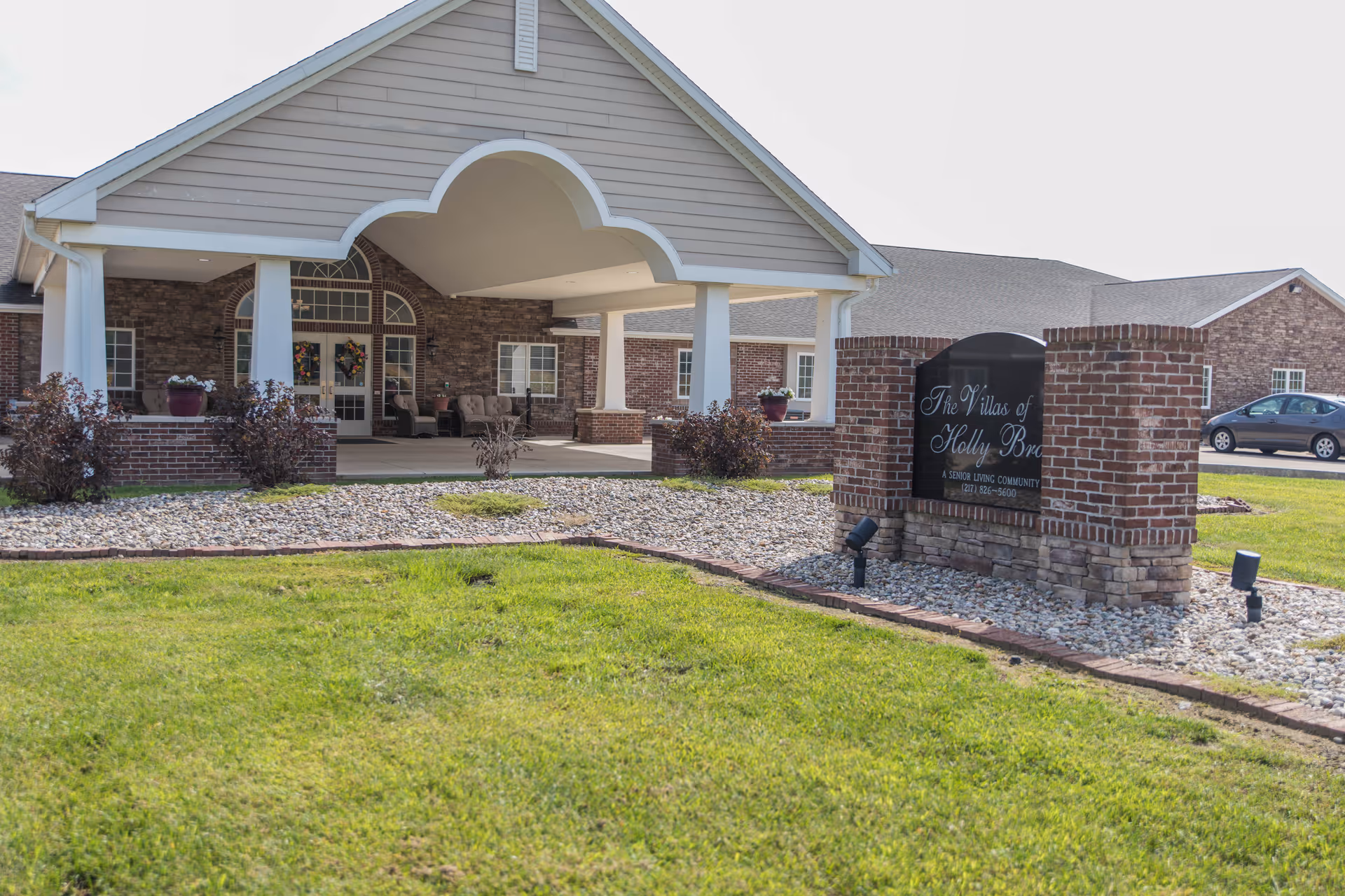 Exterior view of the Villas of Holly Brook Marshall senior living community building with a covered entrance, brick and stone facade, and a landscaped area with grass and decorative rocks. A sign in front displays the community name and contact number.