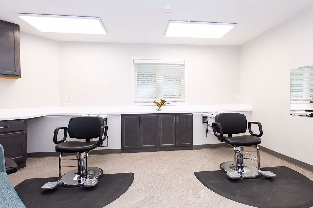 A clean and modern salon area with two black salon chairs on black mats, positioned in front of a white countertop with dark cabinets underneath. A small window with blinds and a vase with flowers is centered on the back wall. The room has bright overhead lighting and light-colored flooring.