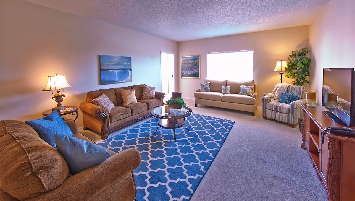A cozy living room with two brown sofas, a beige sofa, a striped armchair, a blue patterned rug, a round glass coffee table, two table lamps, a floor lamp, framed artwork on the walls, and a large window letting in natural light.