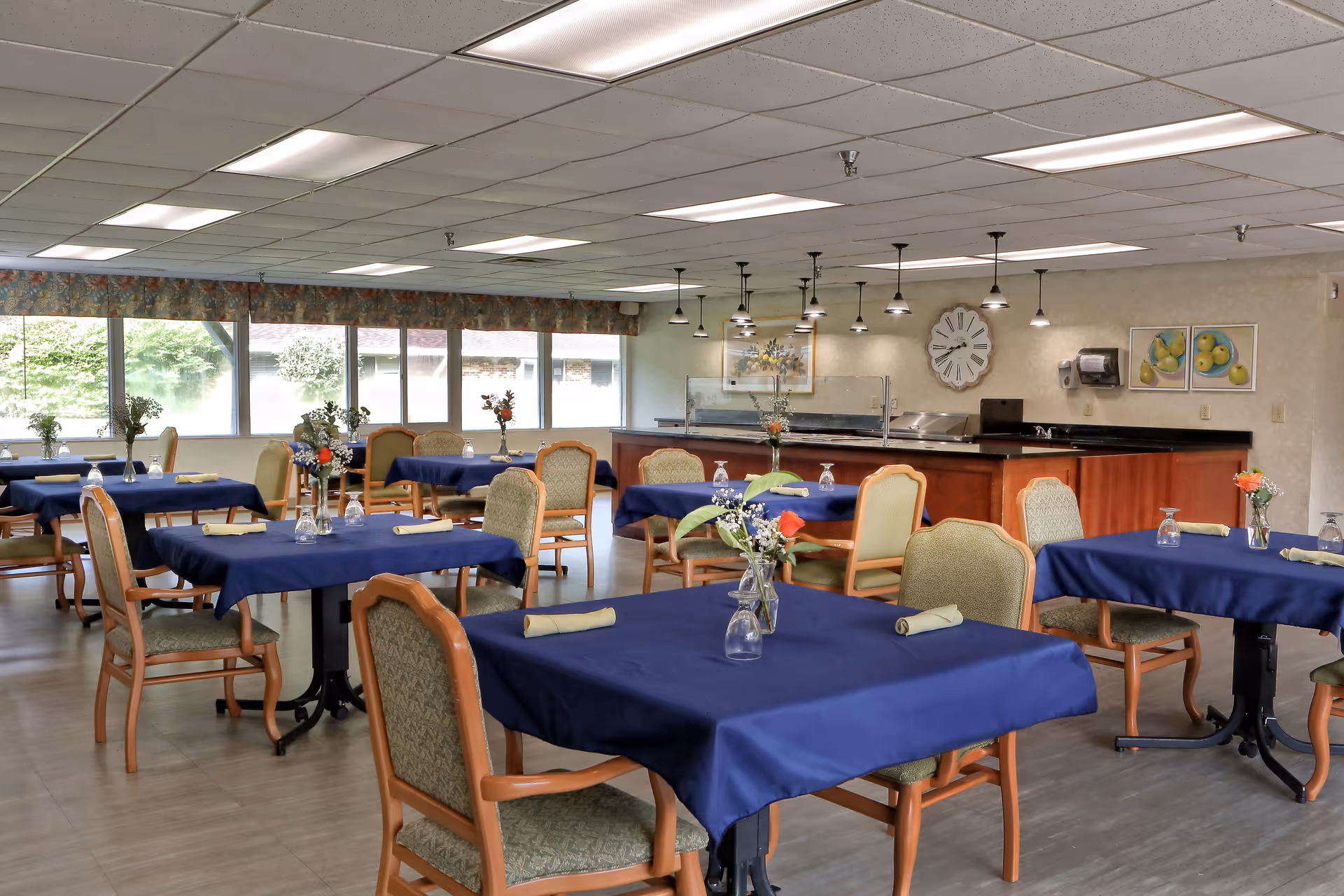 A dining room with multiple tables covered in blue tablecloths, each set with a small vase of flowers and rolled napkins. The room has large windows letting in natural light, cushioned chairs around the tables, and a serving counter with pendant lights hanging above it. The walls are decorated with framed pictures and a large clock.