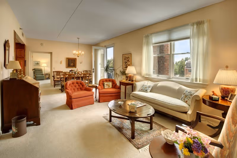 A cozy living room with a beige sofa, two orange armchairs, a wooden coffee table on a small rug, and a side table with a lamp and flowers. In the background, there is a dining area with a wooden dining table and chairs, a chandelier, and a door leading outside. The room has large windows with sheer curtains letting in natural light.