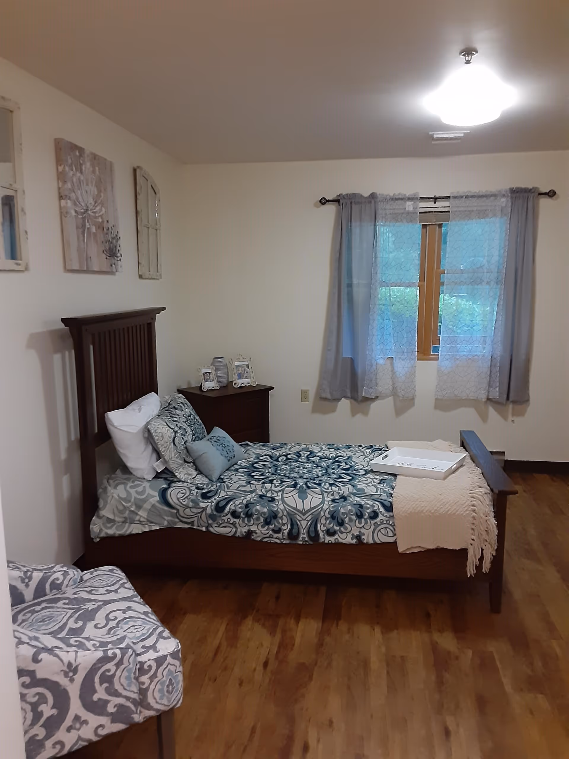 A bedroom with a wooden bed frame and a blue patterned bedspread. There are several pillows on the bed and a white tray placed on top of a beige throw blanket at the foot of the bed. A wooden nightstand with framed photos and a decorative vase is next to the bed. The room has wooden flooring, a window with sheer and solid curtains, and light-colored walls adorned with three pieces of wall art.