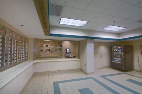 Interior view of a senior living facility lobby or common area with a tiled floor, a vending machine on the right, a service window with shelves and decorative items in the center, and a mail or message slot area on the left wall.