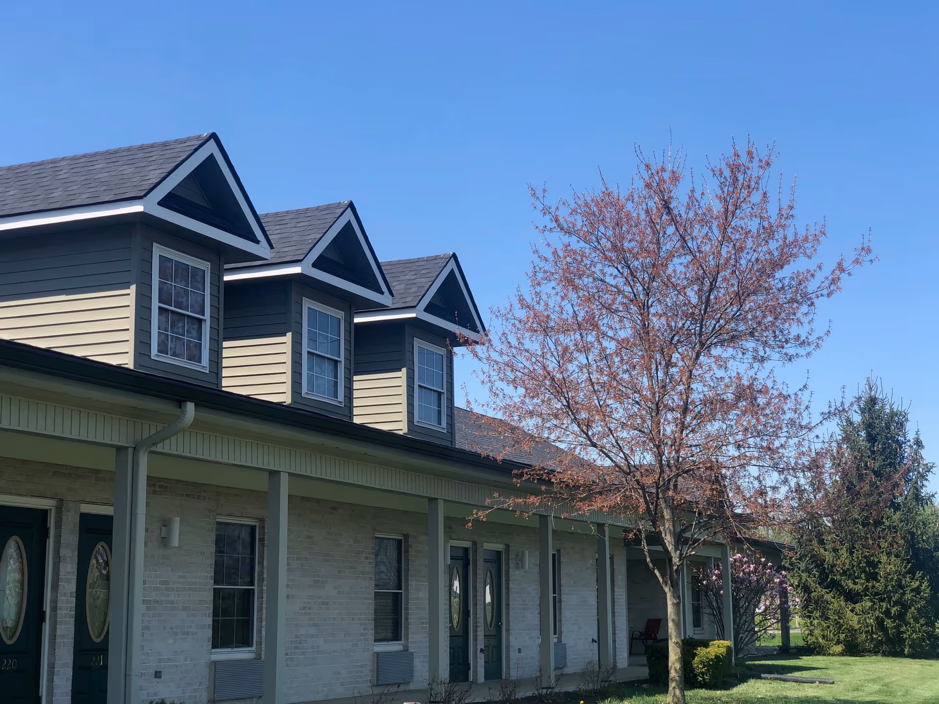 Exterior view of a senior living facility building with beige brick walls, green doors, and three dormer windows on the roof. A tree with budding pink leaves stands in front of the building under a clear blue sky.