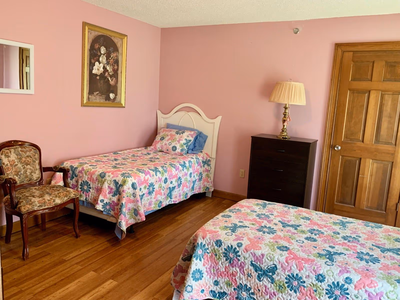 A bedroom with pink walls featuring two twin beds with floral and butterfly patterned bedspreads. There is a wooden chair with floral upholstery, a dark wooden dresser with a brass lamp on top, a wooden door, and a framed floral painting on the wall.