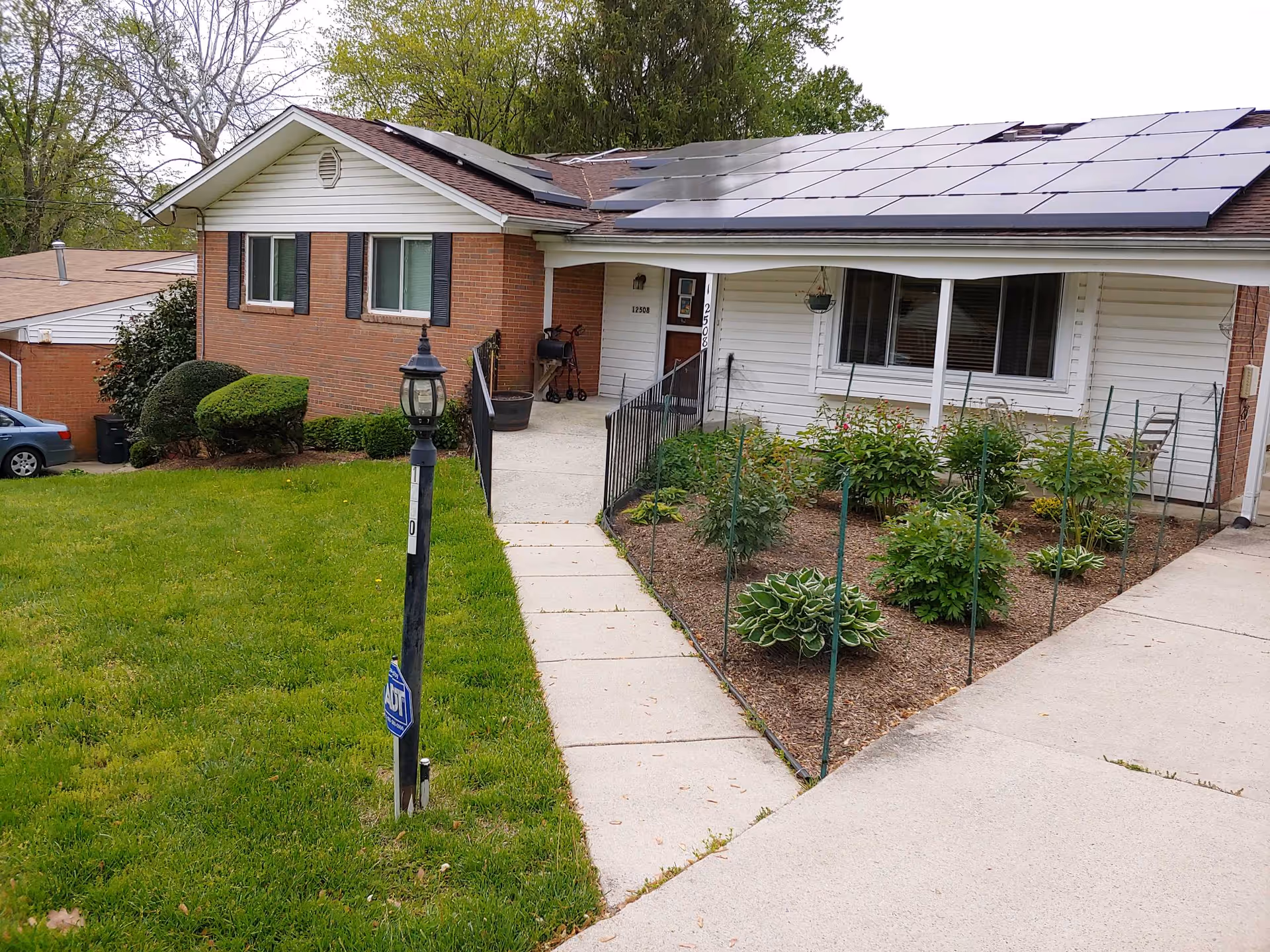 Exterior view of a single-story assisted living facility building with a brick and white siding facade. There is a concrete walkway leading to the entrance, a garden area with plants on the right, and a grassy lawn on the left. Solar panels are visible on the roof, and a lamp post with an ADT security sign is in the front yard.