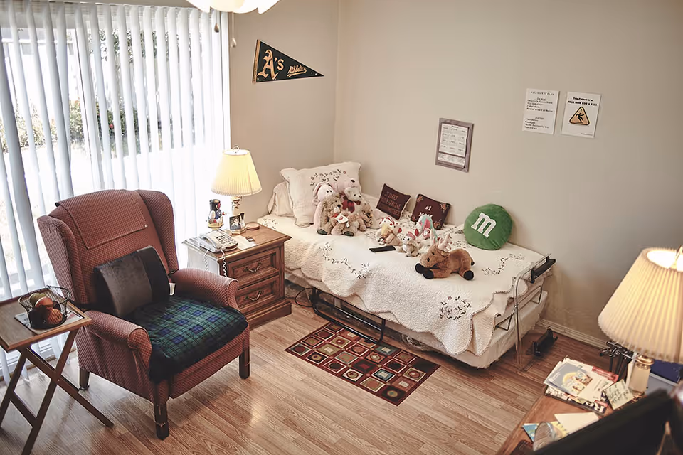 A cozy bedroom in Abigail's Beckham Care Home featuring a single bed with a white quilt and several stuffed animals arranged on it, including teddy bears and a green M&M pillow. Next to the bed is a wooden nightstand with a lamp, a telephone, and some small items. A comfortable armchair with a plaid cushion and a small side table with a fruit basket are positioned near vertical blinds covering a window or sliding door. The room has light-colored walls with a few framed notices and a pennant on the wall, and a small patterned rug on the wooden floor.