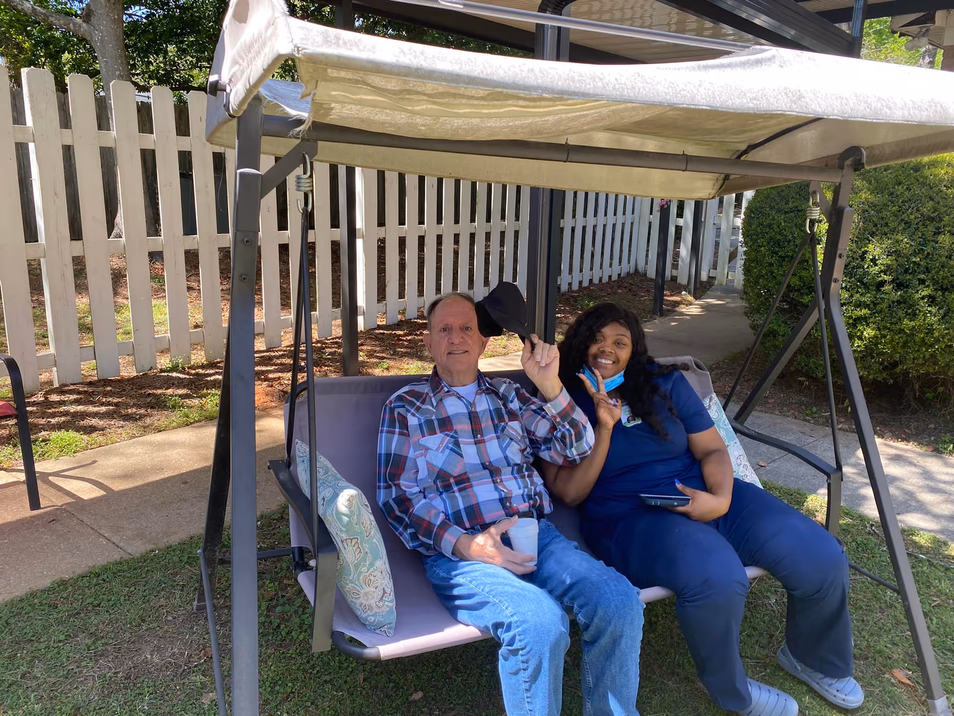 An elderly man and a woman sitting together on an outdoor swing bench with a canopy. The man is wearing a plaid shirt and jeans, holding a cup and tipping his hat, while the woman in blue scrubs is smiling and making a peace sign. They are in a garden area with a white picket fence and greenery in the background.