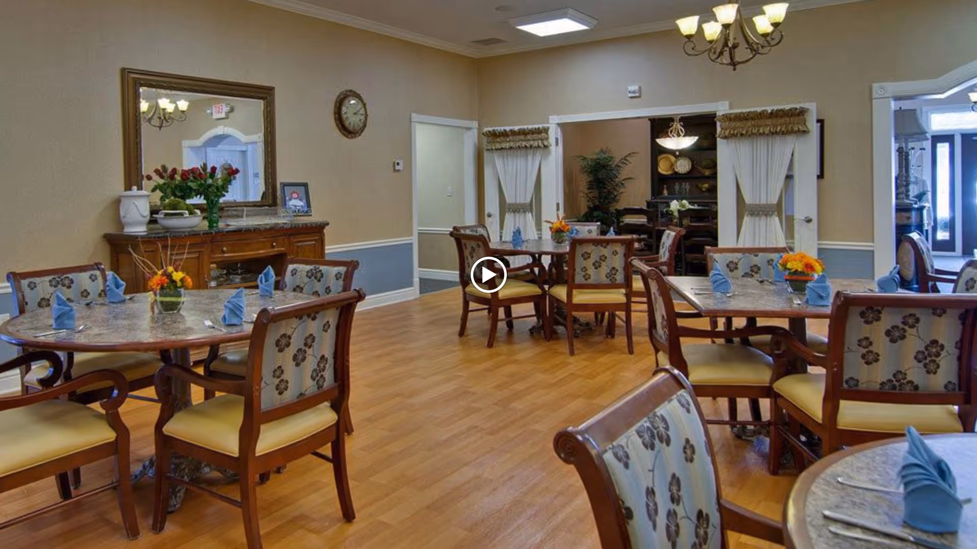 A dining room in a senior living facility with round tables set with blue napkins and floral centerpieces. The room has wooden chairs with floral upholstery, a wooden sideboard with a mirror above it, a wall clock, and warm beige walls with white trim. There is a chandelier hanging from the ceiling and an open doorway leading to another room.