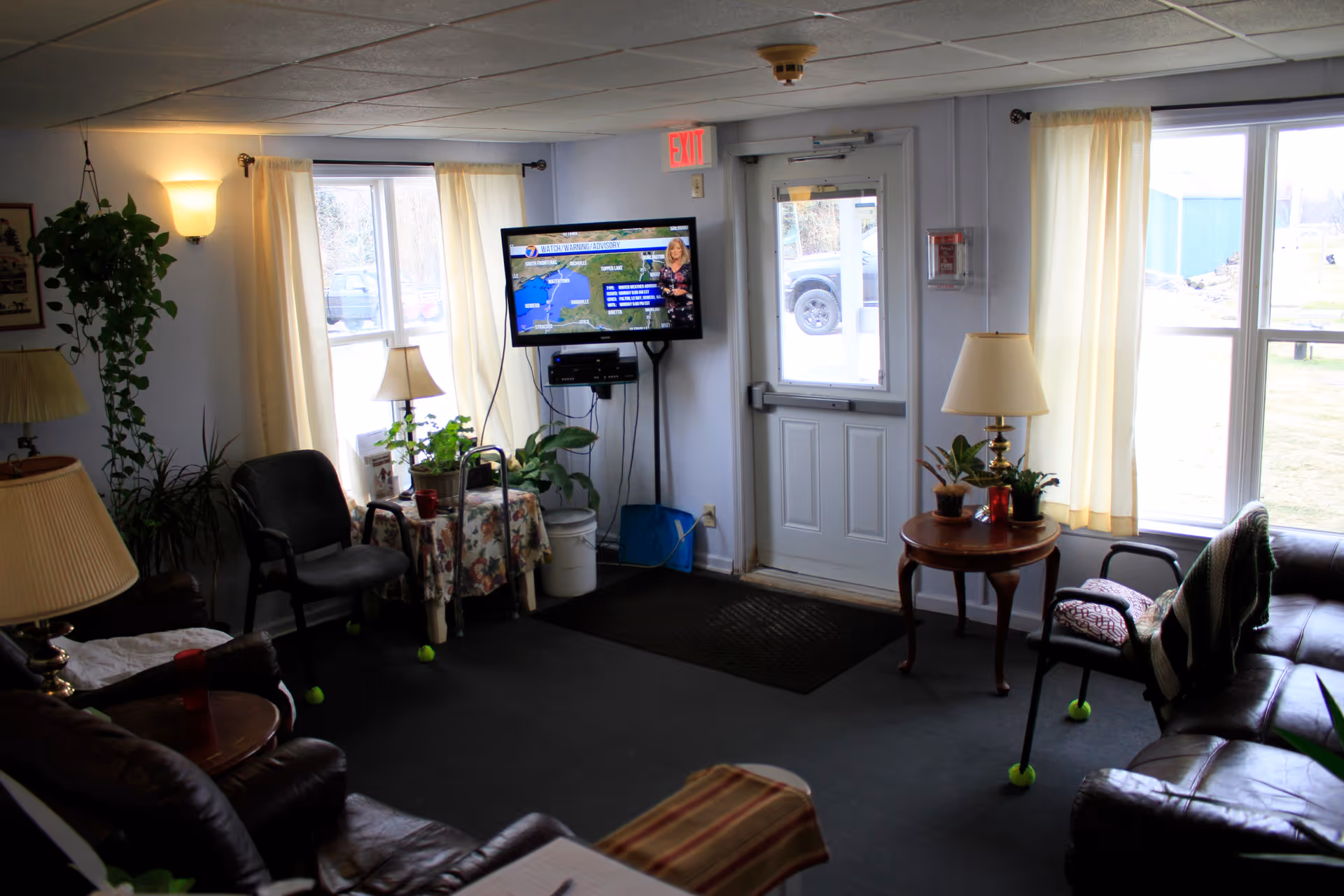 A cozy living room area with several chairs and a sofa arranged around small tables with lamps and potted plants. A flat-screen TV mounted on the wall displays a weather forecast. Large windows with light curtains let in natural light, and a door with a window leads outside. The room has a carpeted floor and soft lighting from wall sconces.