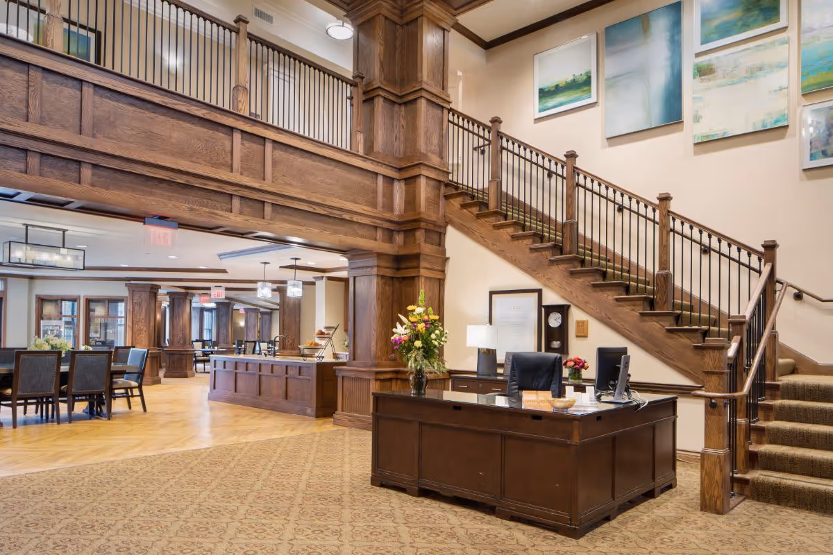 Interior view of a senior living facility lobby with a wooden reception desk, a computer, a vase with flowers, and a staircase with wooden railings. The area features wooden columns and beams, carpeted flooring near the desk, and hardwood flooring in the dining area with tables and chairs. Several framed abstract paintings hang on the wall above the staircase.