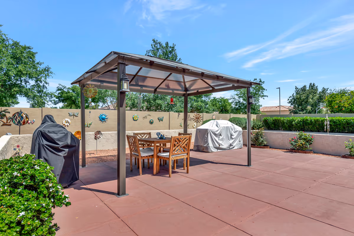 Outdoor patio area with a metal gazebo covering a wooden table and four chairs. Two covered grills are positioned on either side of the gazebo. The surrounding area has decorative wall art, bushes, and trees under a clear blue sky.