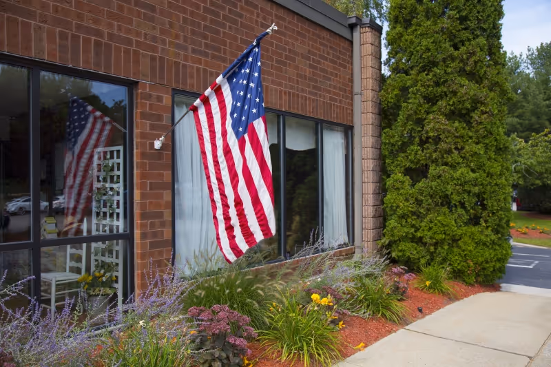 Exterior view of a brick building with an American flag mounted on the wall. There are large windows reflecting the flag and some greenery inside. In front of the building, there is a landscaped garden with various plants and flowers, and a sidewalk runs alongside it. Tall evergreen trees and a parking area are visible in the background.