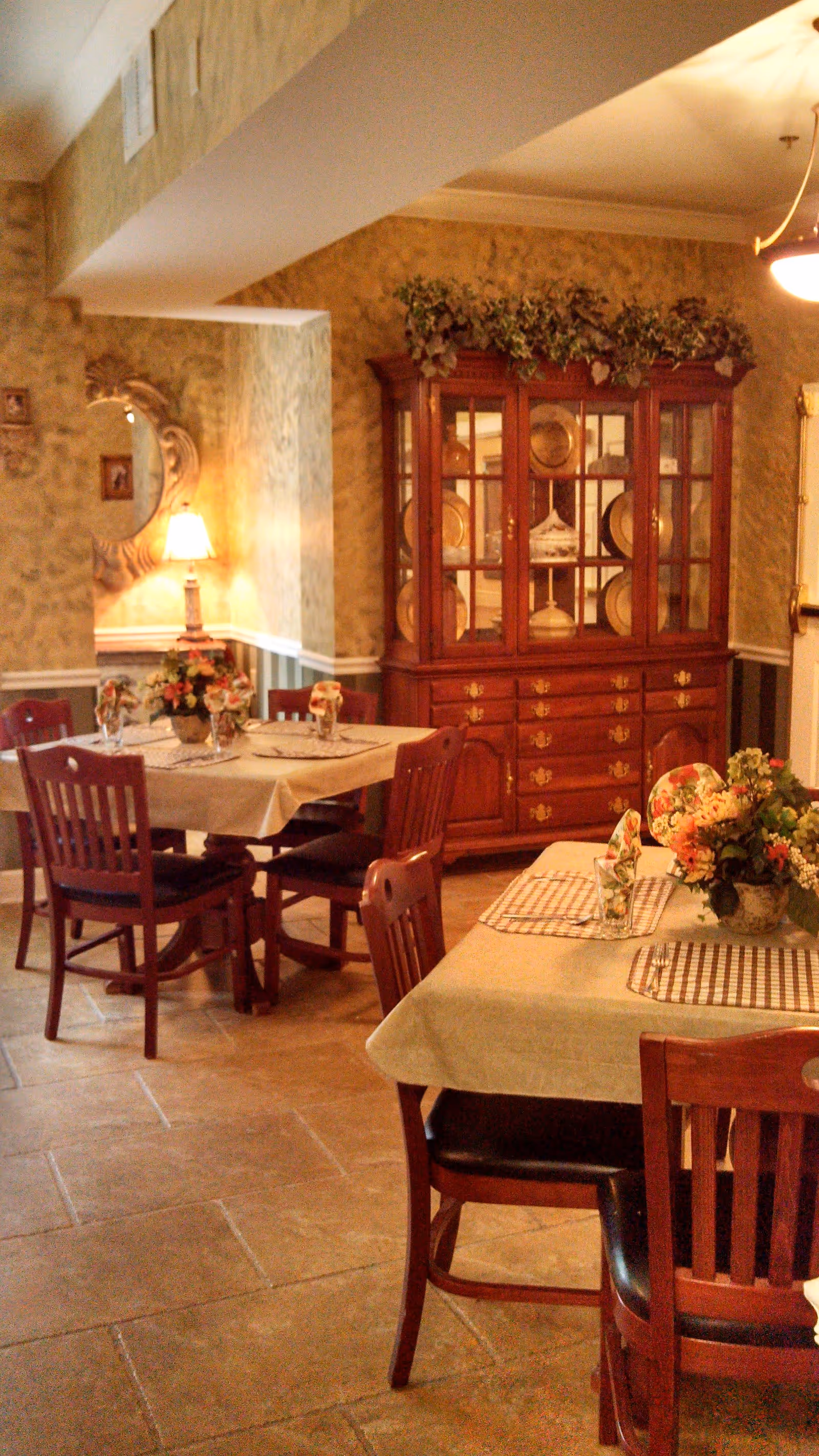 A cozy dining room with two tables covered with beige tablecloths, each set with placemats, napkins, and floral centerpieces. The room features wooden chairs, a wooden china cabinet displaying plates and decorative items, a wall mirror, and a table lamp providing warm lighting. The walls have a textured, warm-toned wallpaper and the floor is tiled.