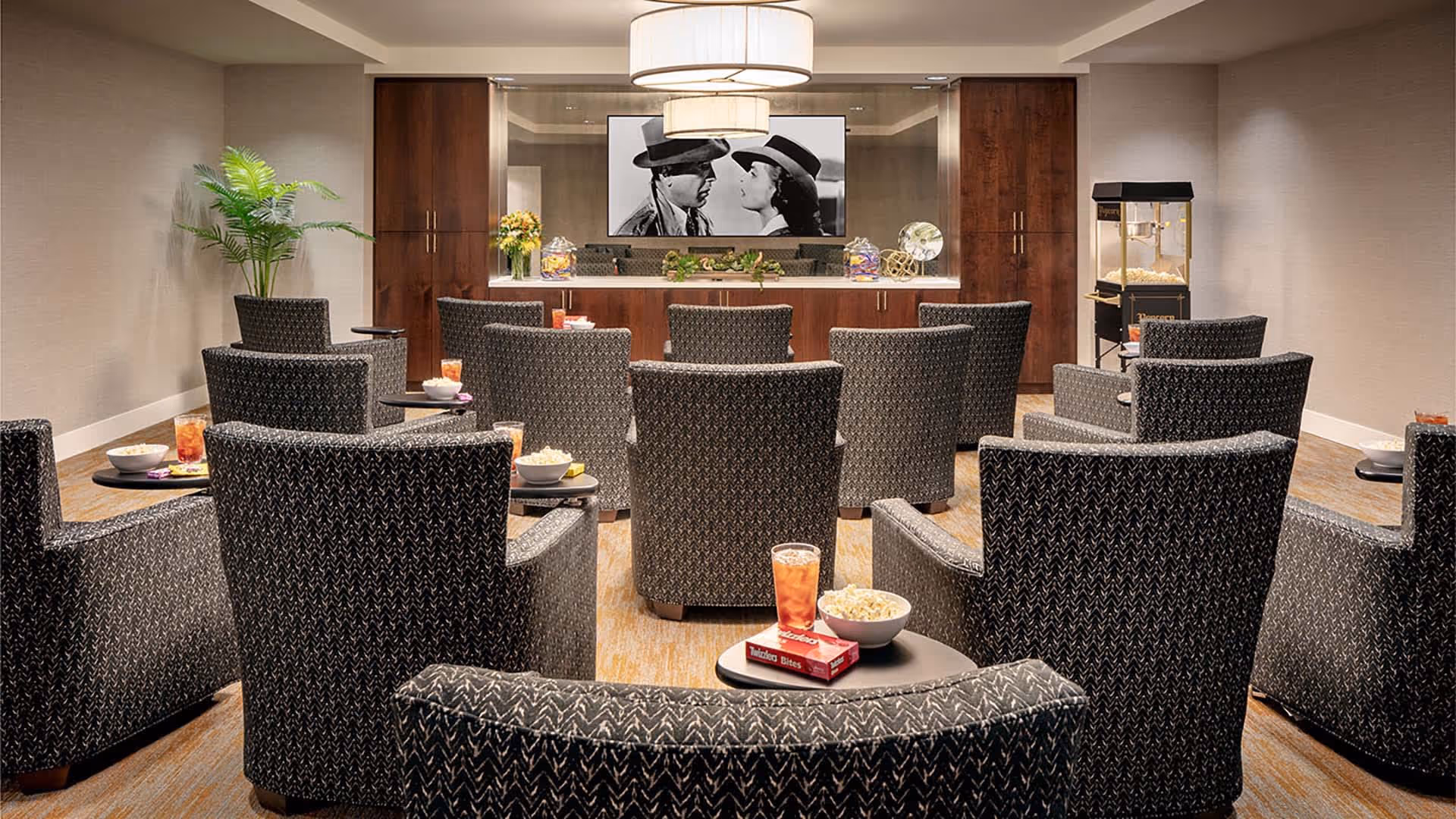 A cozy movie theater room with multiple rows of patterned armchairs facing a large screen showing a black and white movie scene. Each chair has a small side table with a bowl of popcorn and a drink. There is a popcorn machine on the right side and a decorative plant on the left. The room has warm lighting and wooden cabinetry behind the screen.