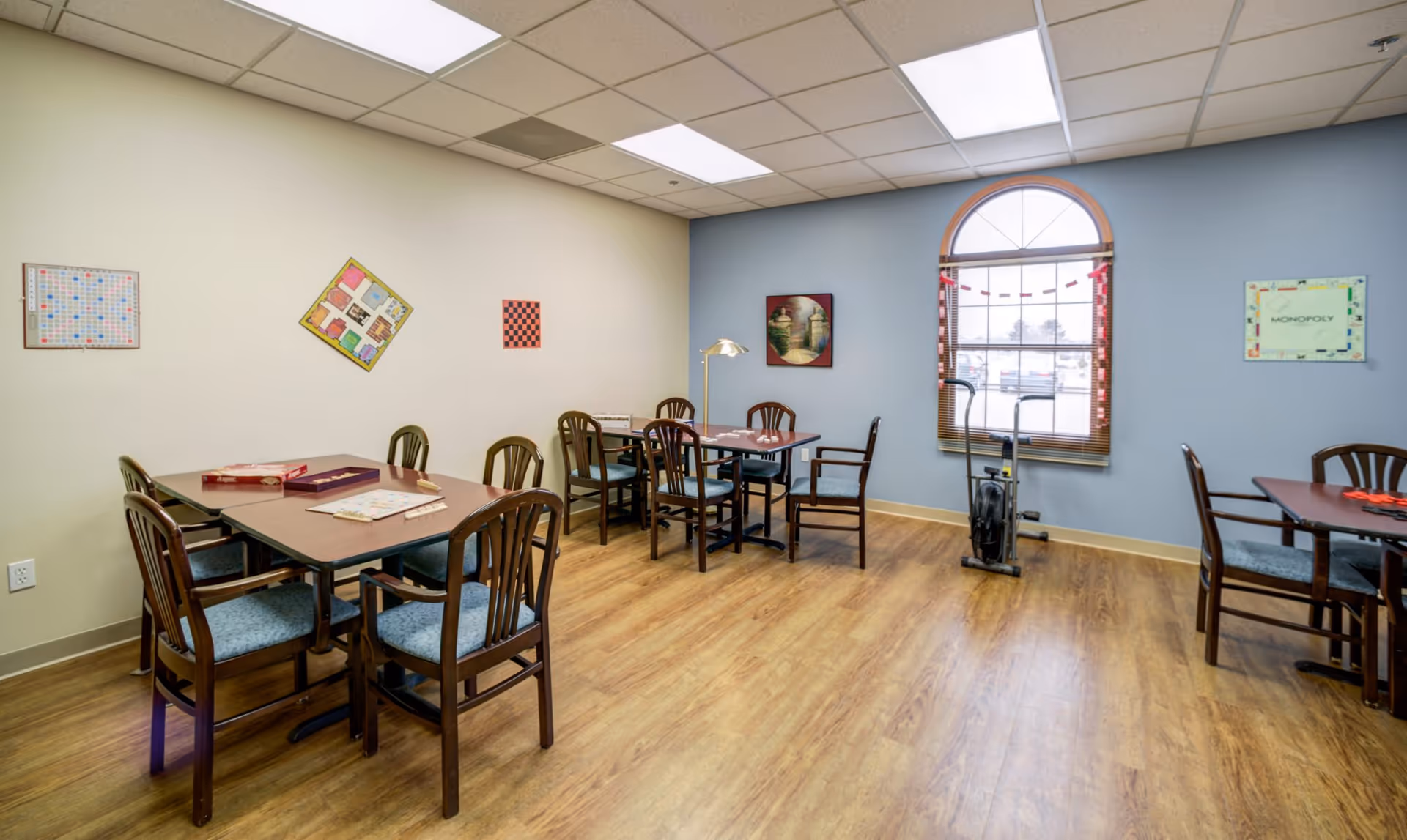 A well-lit activity room with wooden floors and several tables and chairs arranged for group games. The walls are decorated with framed board game boards including Scrabble, Monopoly, and checkers. There is a large arched window with blinds and a paper chain decoration. An exercise bike is positioned near the window.