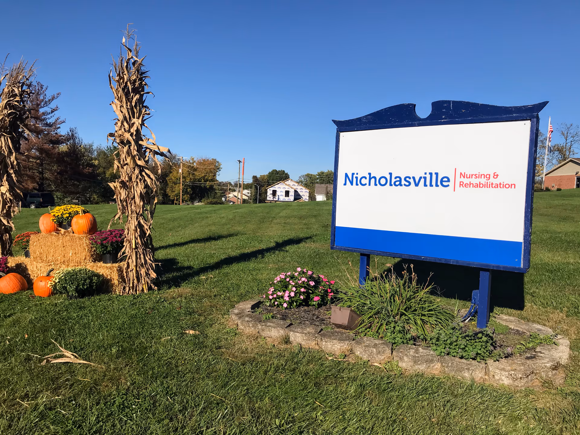 Outdoor scene showing a large sign for Nicholasville Nursing & Rehabilitation on a grassy lawn with a clear blue sky. To the left of the sign, there is a fall-themed decoration with hay bales, pumpkins, flowers, and dried cornstalks. Trees and houses are visible in the background.