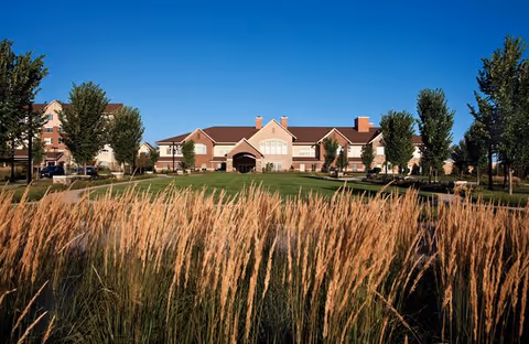 View of a senior living community building with a large lawn and tall grasses in the foreground under a clear blue sky.