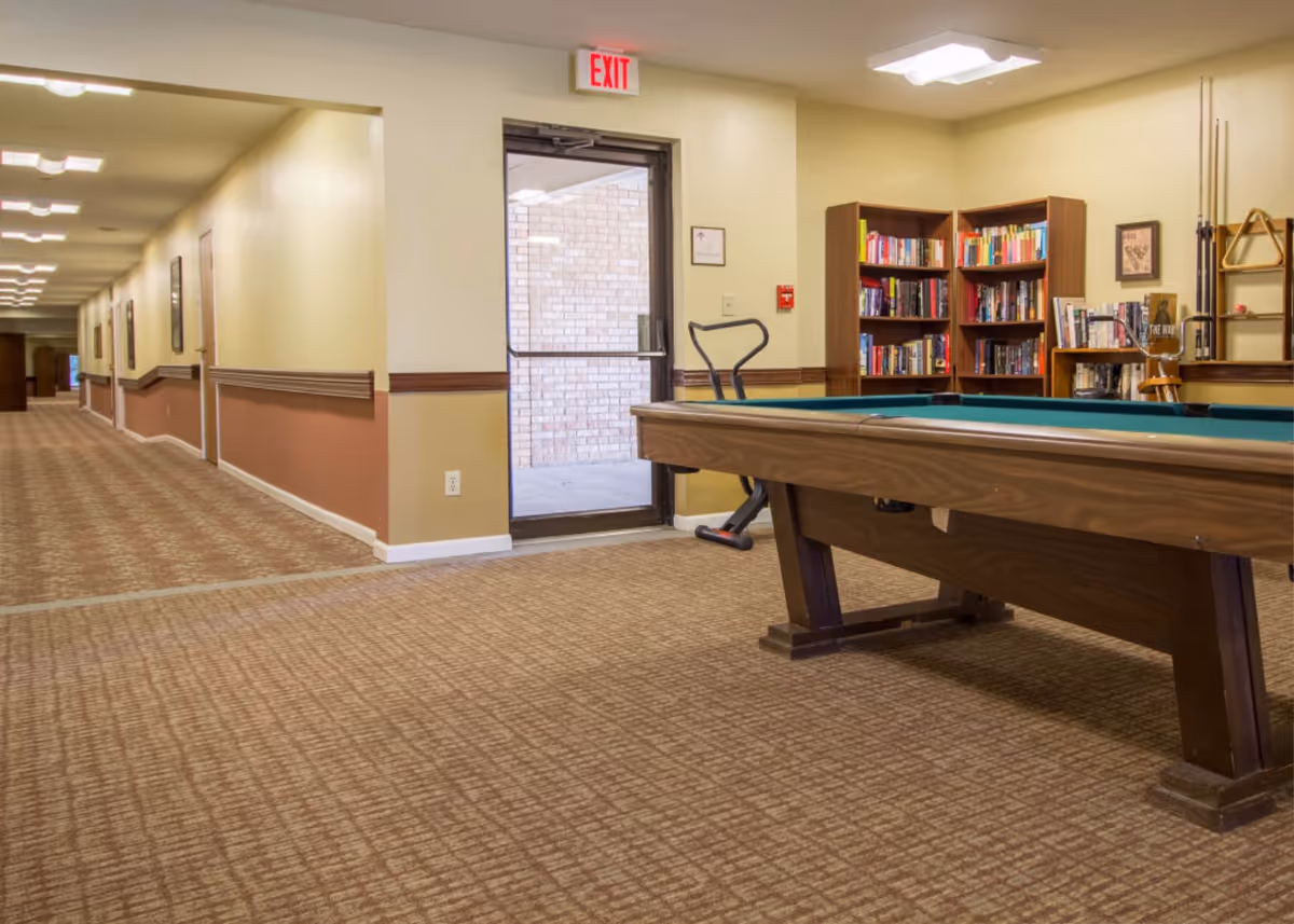 A senior living common area with a pool table, bookshelves, an exercise bike and a long carpeted hallway leading to an exit door.