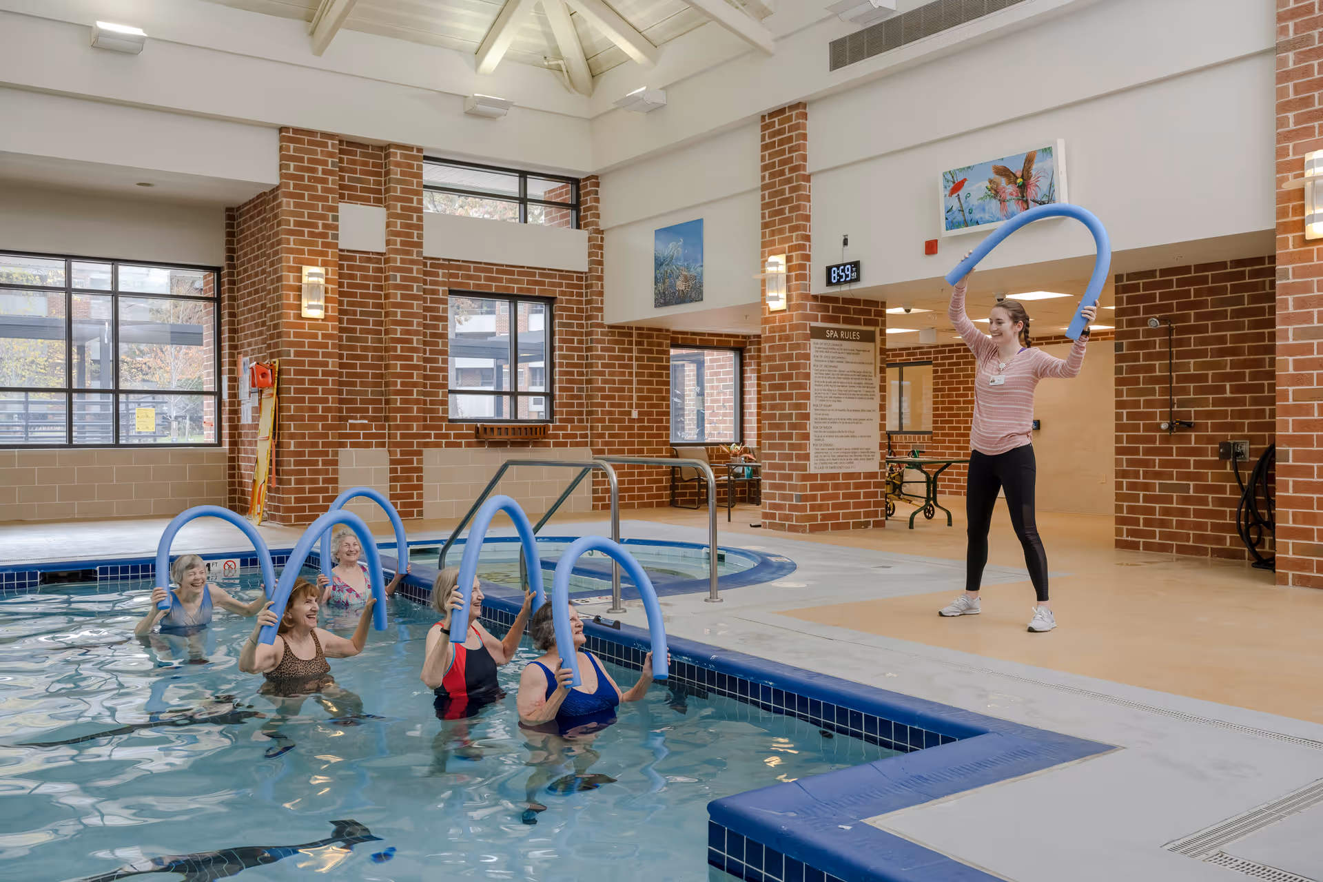 A group of elderly women in a swimming pool participating in a water exercise class led by a young female instructor holding blue pool noodles. The indoor pool area features brick walls, large windows, and a high ceiling with exposed beams.
