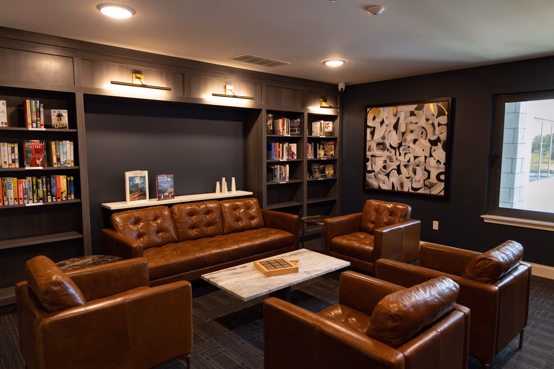 A cozy living room area with a brown leather sofa and four matching leather armchairs arranged around a rectangular marble coffee table. Behind the sofa are built-in dark wood bookshelves filled with books and decorative items. A large abstract painting hangs on the dark wall to the right, and a window lets in natural light.