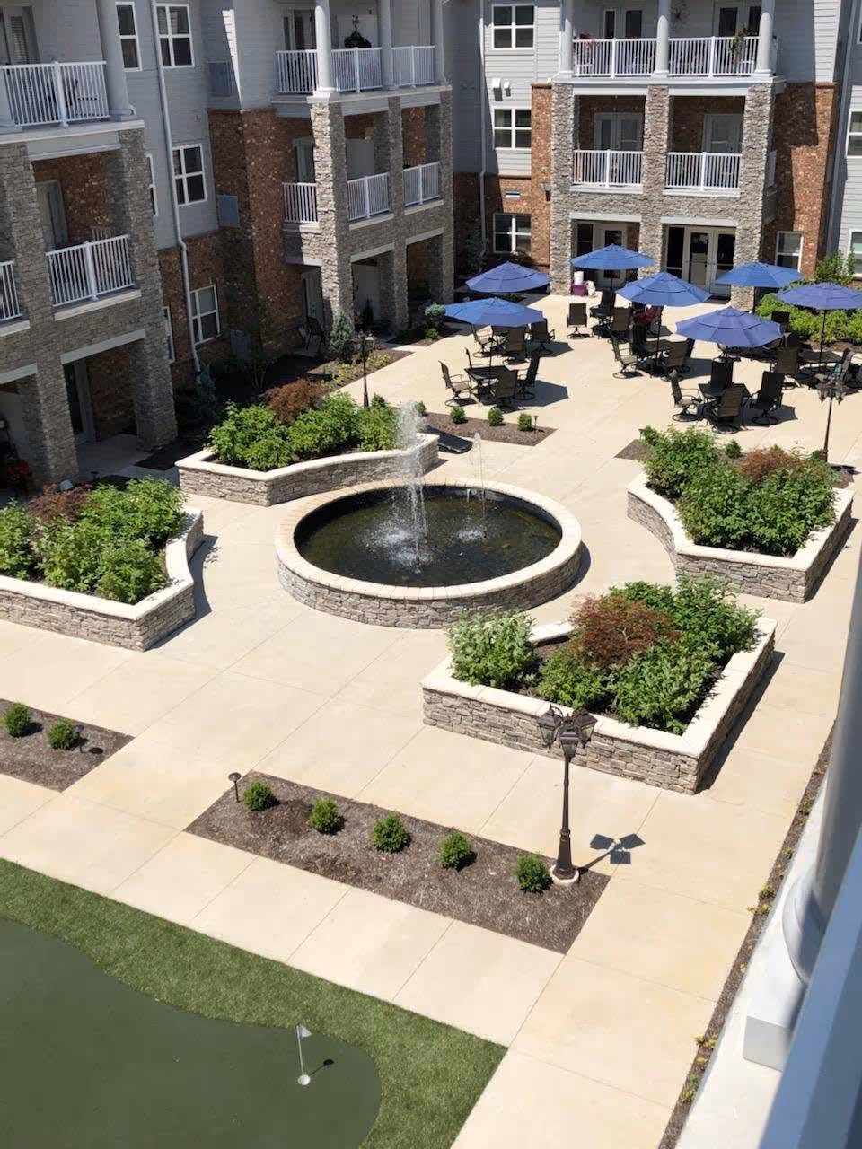 Outdoor courtyard area of a senior living facility with a central circular water fountain surrounded by raised stone planters filled with greenery. Several tables with blue umbrellas and chairs are arranged on the paved patio area. The multi-story building with balconies and large windows surrounds the courtyard. A small putting green with a flag is visible in the foreground.